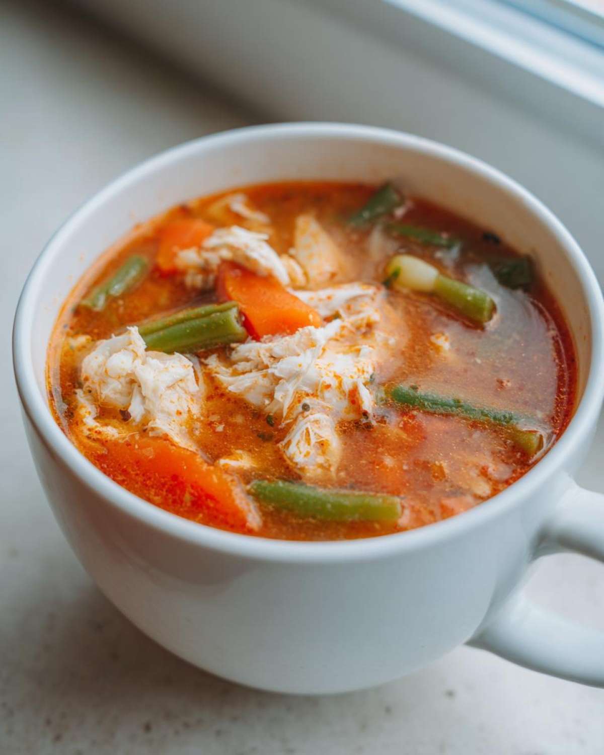 Close-up of a white mug filled with rich, tomato-based Maryland crab soup, featuring lump crab meat, carrots, and green beans.