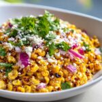 Close-up of a white bowl filled with charred corn kernels, topped with crumbled white cheese, red onion, and fresh cilantro for Mexican street corn salad.
