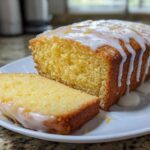 A moist Lemon Loaf cake, partially sliced, covered in a thick white lemon glaze and zest.