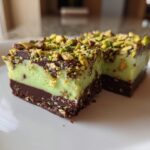 Two rich, layered No-Bake Pistachio Chocolate Fudge Squares topped with chopped pistachios, served on a white plate.