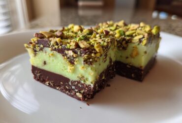 Two rich, layered No-Bake Pistachio Chocolate Fudge Squares topped with chopped pistachios, served on a white plate.
