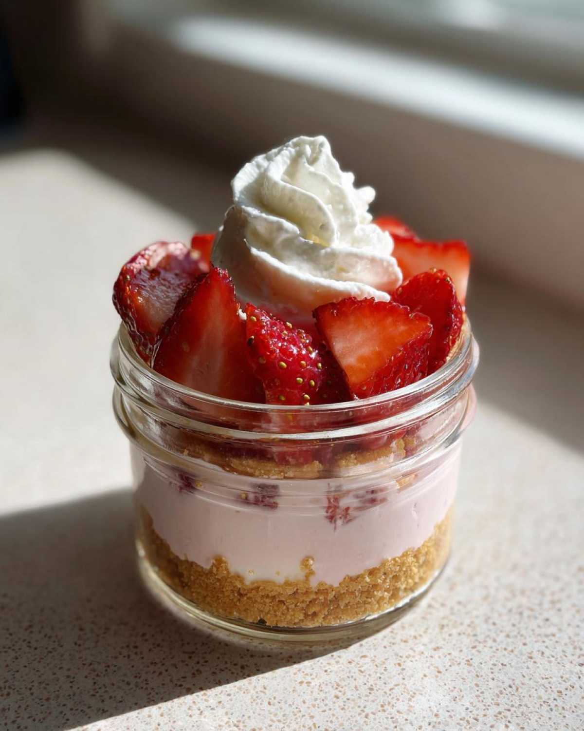 A single serving of No-Bake Strawberry Cream Pie Jars layered with graham cracker crust, pink cream, and topped with fresh strawberries and whipped cream.