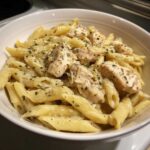 A close-up of creamy chicken pasta made with penne noodles, topped with seasoned chicken pieces and herbs.