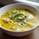 A close-up of a white bowl filled with One-Pot Creamy Lemon Chicken Noodle Soup, topped with fresh parsley.