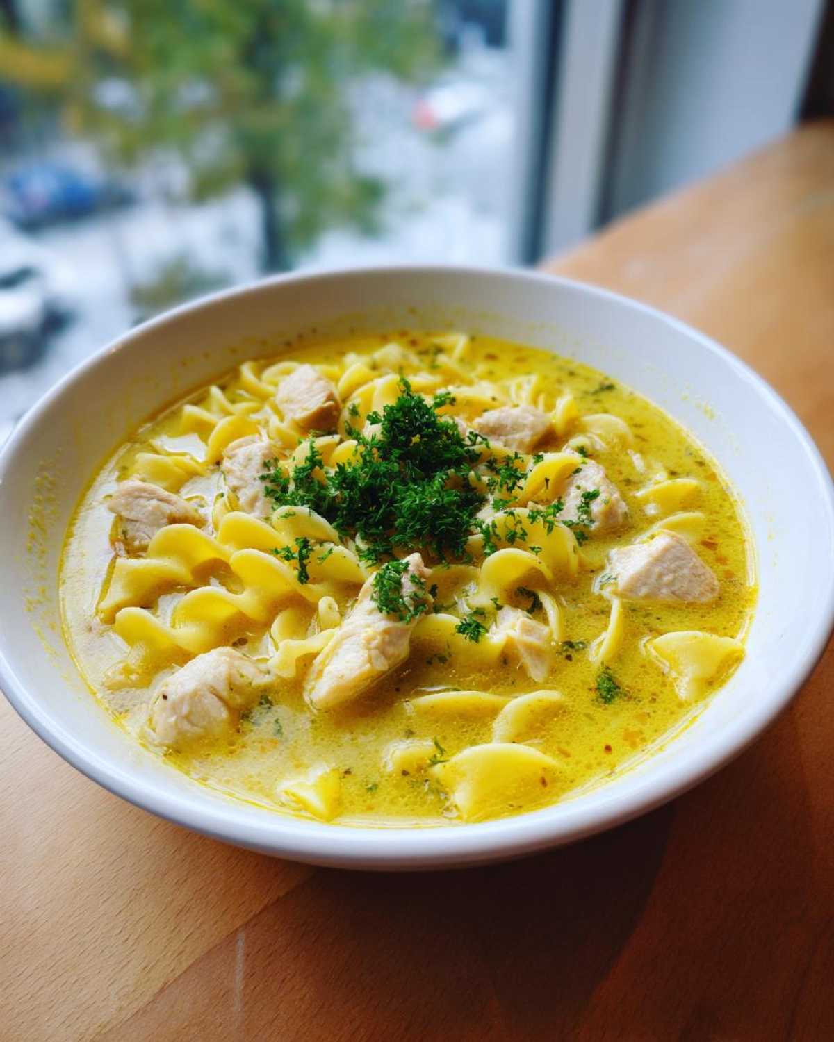 A close-up of a white bowl filled with One-Pot Creamy Lemon Chicken Noodle Soup, garnished with fresh parsley.