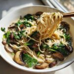 Chopsticks lifting noodles from a bowl of One-Pot Creamy Mushroom Spinach Ramen with rich sauce, mushrooms, and wilted spinach.
