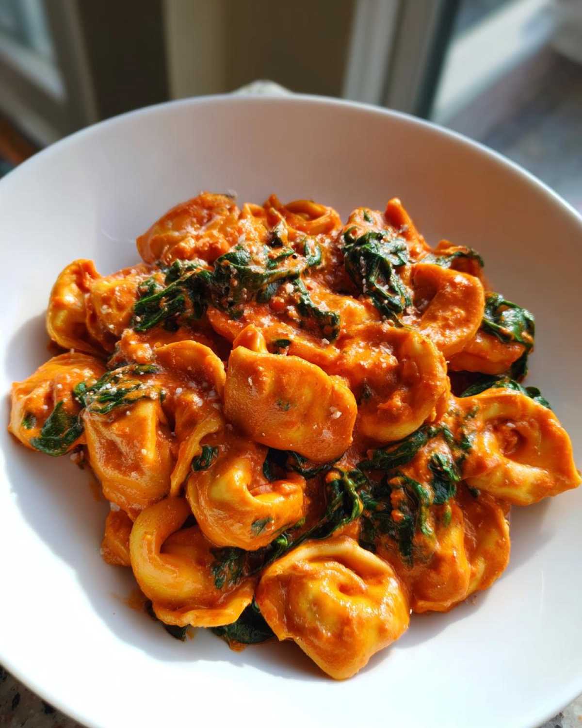 A close-up of One Pot Creamy Tomato Spinach Tortellini coated in a rich orange sauce and mixed with wilted spinach, served in a white bowl.