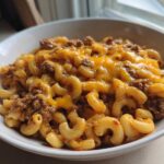 A close-up of a white bowl filled with creamy taco pasta mixed with seasoned ground beef and topped with melted cheddar cheese.