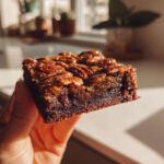 A hand holds up a rich, fudgy square of pecan pie brownies topped with glazed pecans, catching the sunlight.