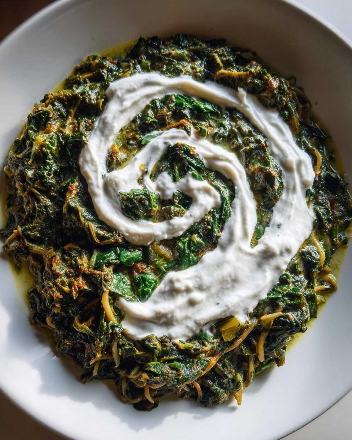 Overhead view of a bowl of rich, dark green Persian noodle soup topped with a swirl of white yogurt or kashk.