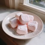 A small plate holding several pink, cube-shaped cream cheese mints dusted heavily with powdered sugar.