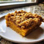 A square slice of rich, orange pumpkin crisp with a thick, crumbly oat topping served on a white plate.