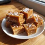 A pile of rich, golden brown salted caramel fudge squares topped with coarse sea salt flakes.