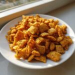 A close-up of a white plate piled high with Savory Garlic Butter Chex Mix Crunchy Snack, seasoned with visible black pepper.