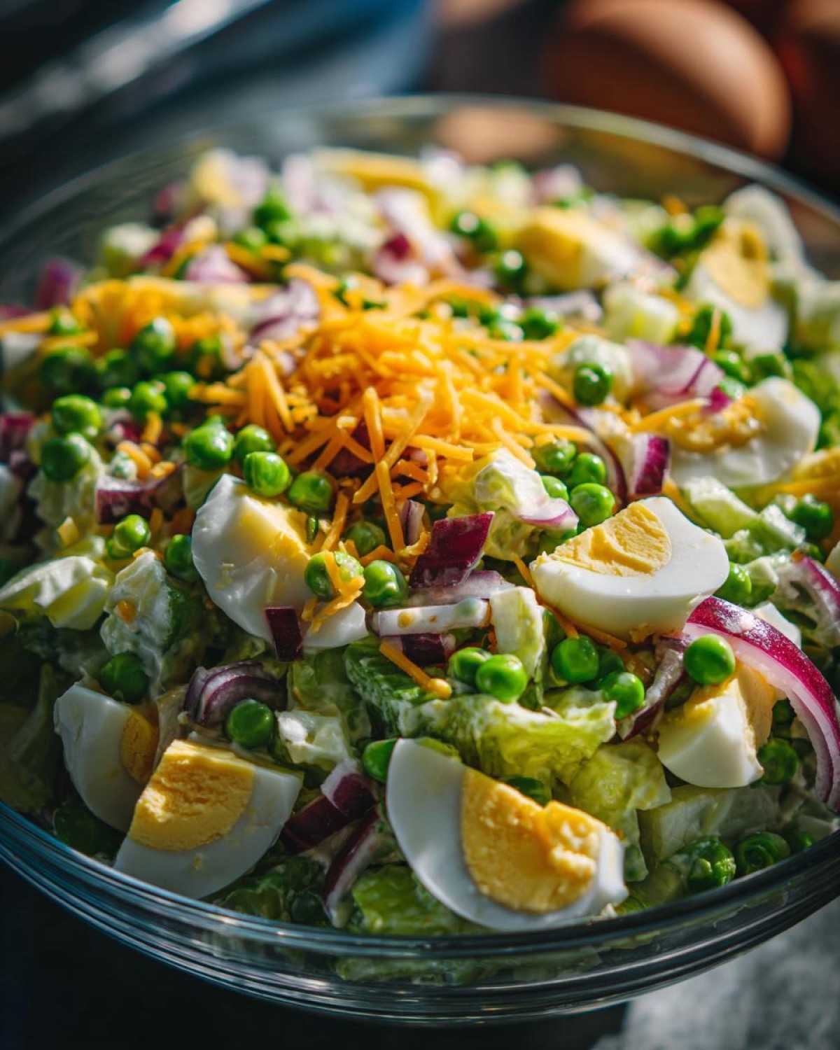 A close-up view of the top layer of a seven layer salad, featuring hard-boiled egg wedges, green peas, shredded cheddar, and red onion.
