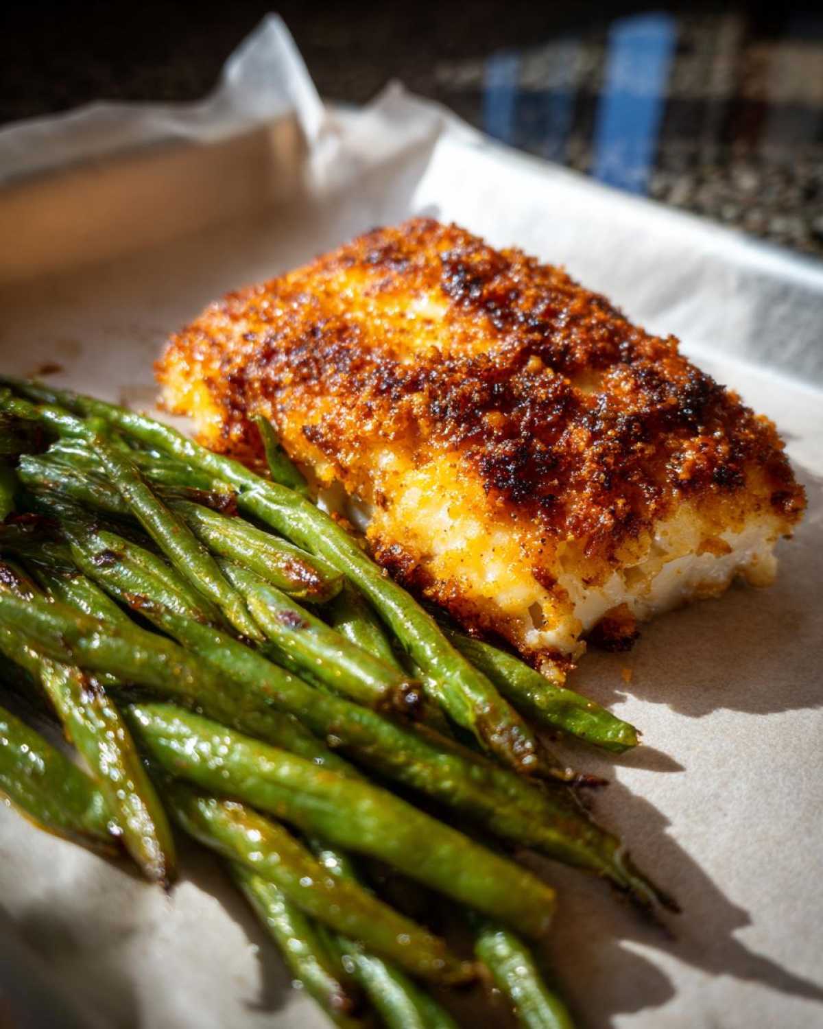 A close-up of Sheet Pan Crispy Parmesan Tilapia next to a pile of roasted green beans on parchment paper.