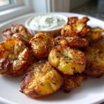 A plate of golden brown Sheet Pan Crispy Smashed Potatoes garnished with herbs, served with a small bowl of garlic aioli.