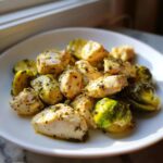 Close-up of cooked chicken pieces and halved Brussels sprouts coated in garlic butter herbs, served on a white plate.