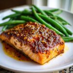 A close-up of a perfectly cooked salmon fillet topped with a thick honey mustard glaze, served next to bright green beans.