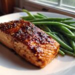 A perfectly cooked fillet of Sheet Pan Honey Mustard Salmon with a thick, glossy glaze next to bright green beans on a white plate.