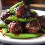 Close-up of glossy Sheet Pan Sticky Sesame Meatballs coated in dark sauce, served with vibrant green snap peas.