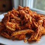 Close-up of vibrant, shredded chicken tinga piled high on a white plate, coated in rich red sauce.