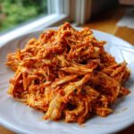 A close-up of shredded chicken tinga piled high on a white plate, coated in a vibrant red, savory sauce.