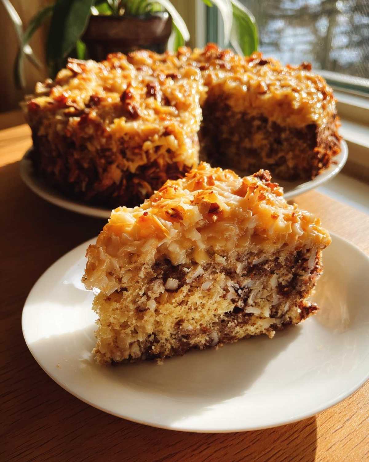 A close-up slice of moist german chocolate poke cake topped with coconut pecan frosting, with the rest of the cake visible in the background.