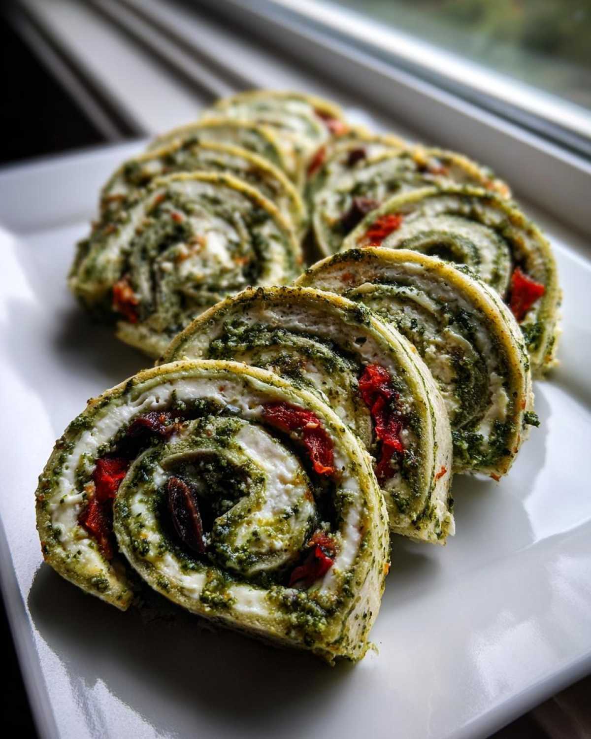 Close-up of several slices of savory italian pinwheels filled with pesto, cream cheese, and sun-dried tomatoes on a white platter.