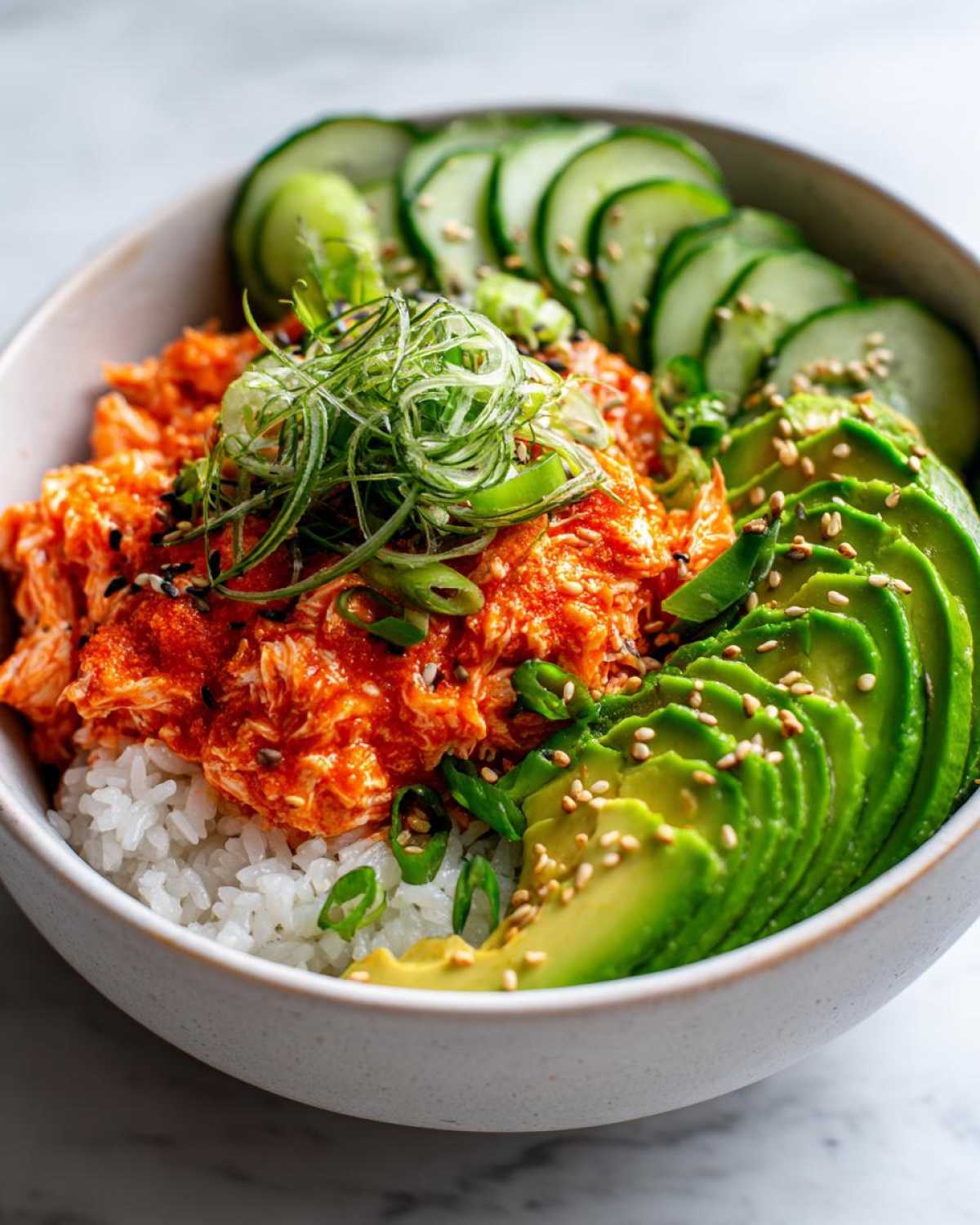 A close-up of a spicy crab sushi bowl featuring rice topped with bright orange spicy crab mix, sliced avocado, cucumber, and scallions.