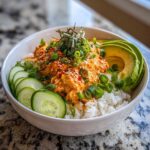 A vibrant spicy crab sushi bowl featuring creamy orange crab mixture over white rice, topped with green onions, sesame seeds, and served with sliced avocado and cucumber.