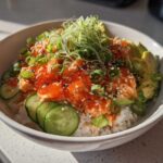 A vibrant spicy crab sushi bowl featuring rice topped with spicy crab mixture, bright orange tobiko, sliced avocado, cucumber, and green onions.