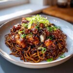 A close-up of Sticky Garlic Chicken Noodles topped with glazed chicken pieces, sesame seeds, and sliced green onions.