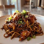 A close-up of Sticky Garlic Chicken Noodles coated in dark sauce, topped with sesame seeds and green onions.