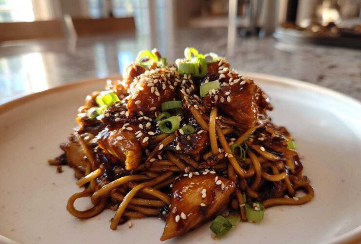 A close-up of Sticky Garlic Chicken Noodles coated in dark sauce, topped with sesame seeds and green onions.