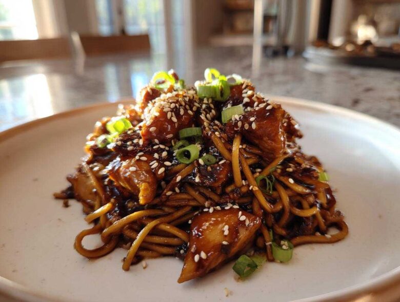 A close-up of Sticky Garlic Chicken Noodles coated in dark sauce, topped with sesame seeds and green onions.