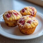 Three golden-brown Strawberry Yogurt Muffins topped with visible strawberry pieces and a white glaze drizzle on a white plate.