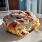 A close-up of a single, fluffy Tres Leches Cinnamon Roll topped with white glaze, sitting on a white plate.