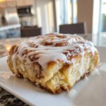 A single, fluffy Tres Leches Cinnamon Roll generously topped with white icing, resting on a white plate.