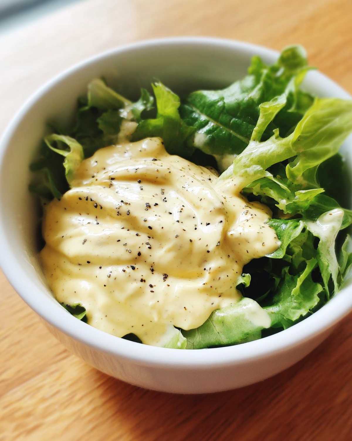 Close-up of a small white bowl filled with fresh green lettuce topped generously with Vegan Creamy Lemon Tahini Dressing and cracked black pepper.