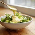 A golden spoon pours thick Vegan Creamy Lemon Tahini Dressing over fresh green lettuce in a white bowl.