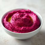 Close-up of vibrant, magenta Vegan Creamy Roasted Beet Hummus swirled in a white bowl, drizzled with olive oil.
