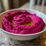 A close-up of bright magenta Vegan Creamy Roasted Beet Hummus swirled in a white bowl, drizzled with olive oil.