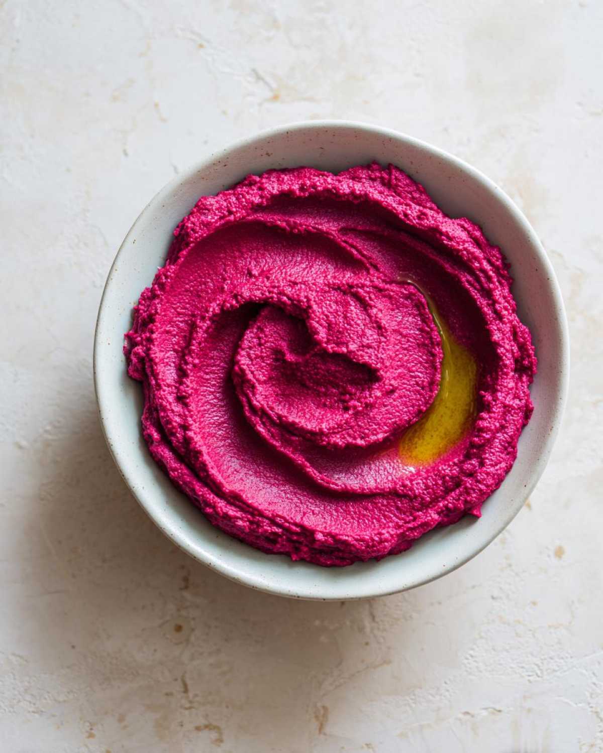 Overhead view of bright magenta Vegan Creamy Roasted Beet Hummus swirled in a light bowl, drizzled with olive oil.