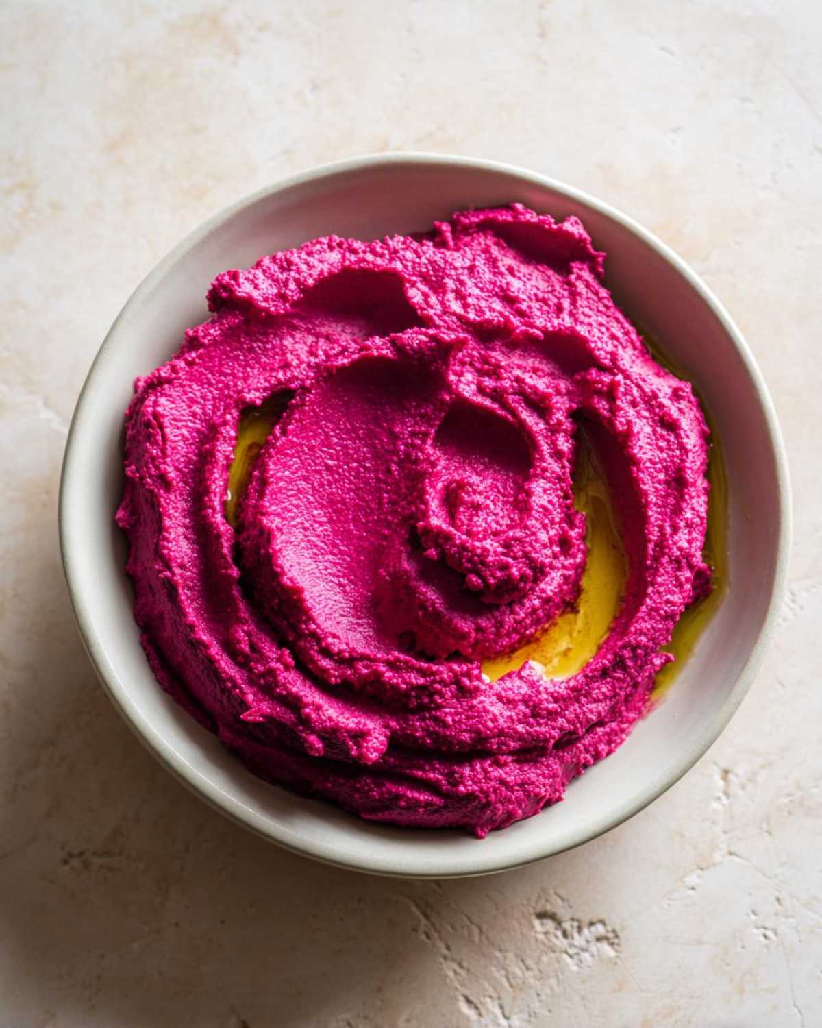 Overhead view of bright pink Vegan Creamy Roasted Beet Hummus swirled in a light gray bowl, drizzled with olive oil.