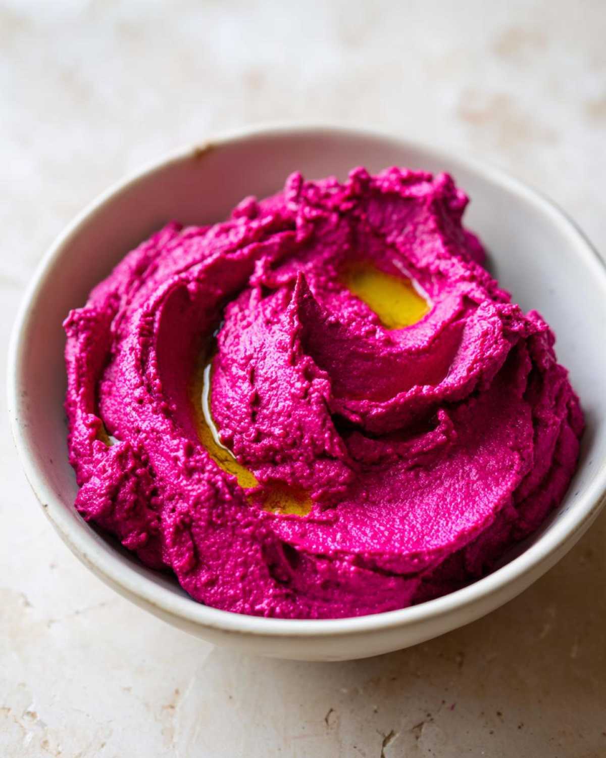 A close-up of vibrant pink Vegan Creamy Roasted Beet Hummus swirled in a white bowl and drizzled with olive oil.