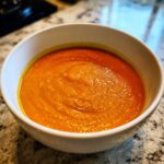 Close-up of a white bowl filled with vibrant orange Vegan Creamy Roasted Carrot Ginger Soup.