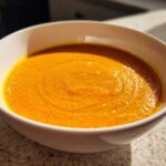 Close-up of a white bowl filled with vibrant orange Vegan Creamy Roasted Carrot Ginger Soup.
