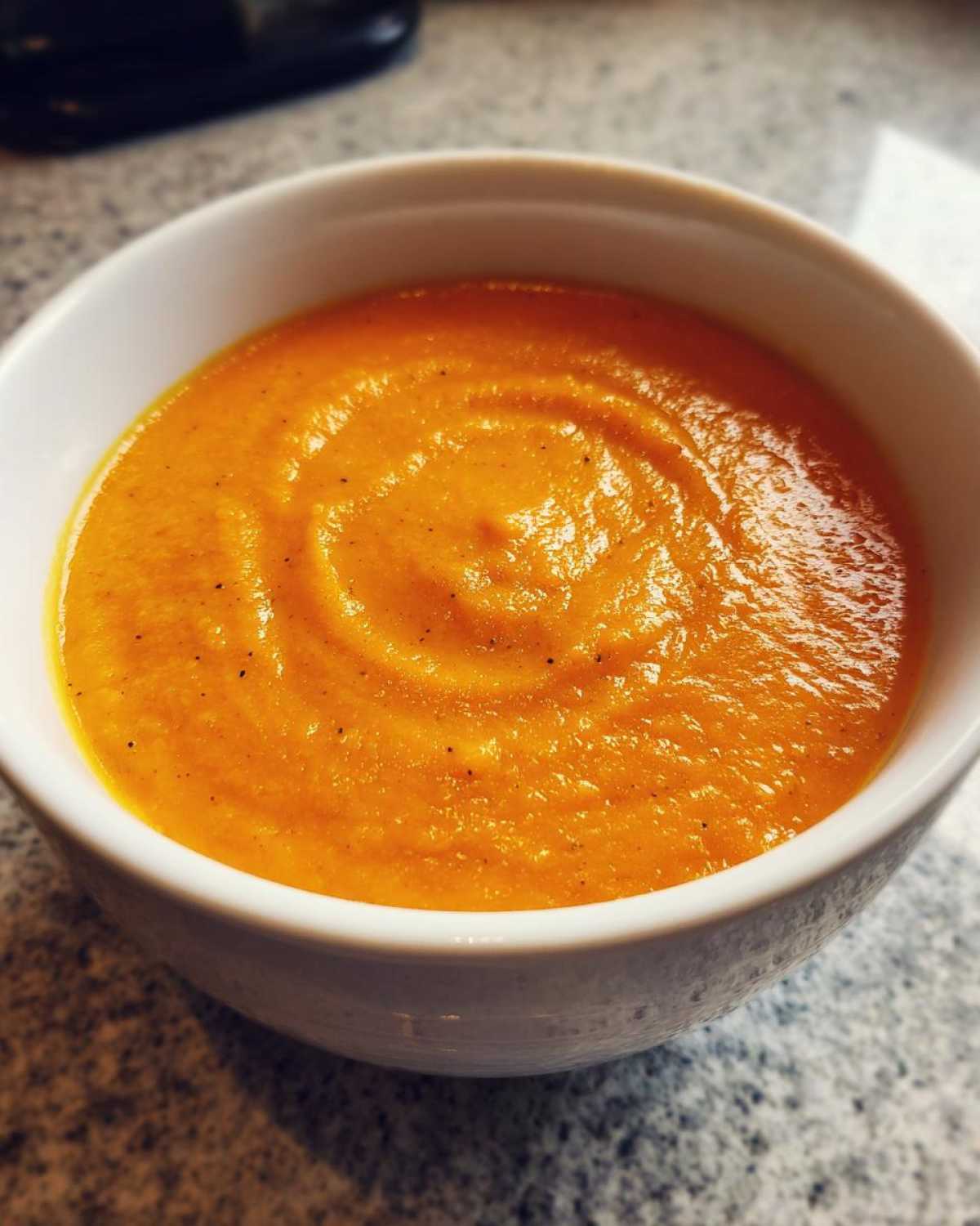 Close-up of a white bowl filled with vibrant orange Vegan Creamy Roasted Carrot Ginger Soup, swirled on top.