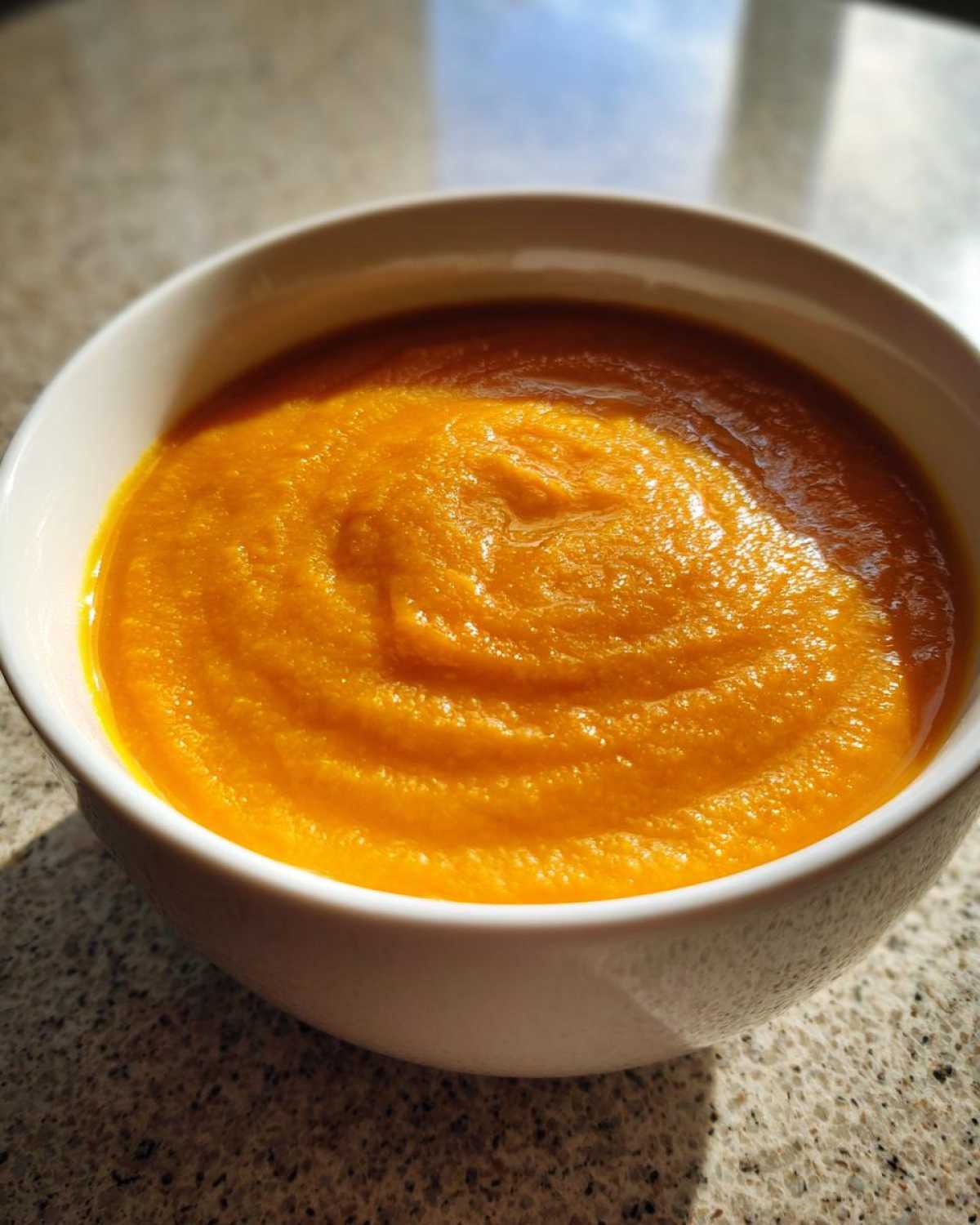 Close-up of a white bowl filled with vibrant orange Vegan Creamy Roasted Carrot Ginger Soup.